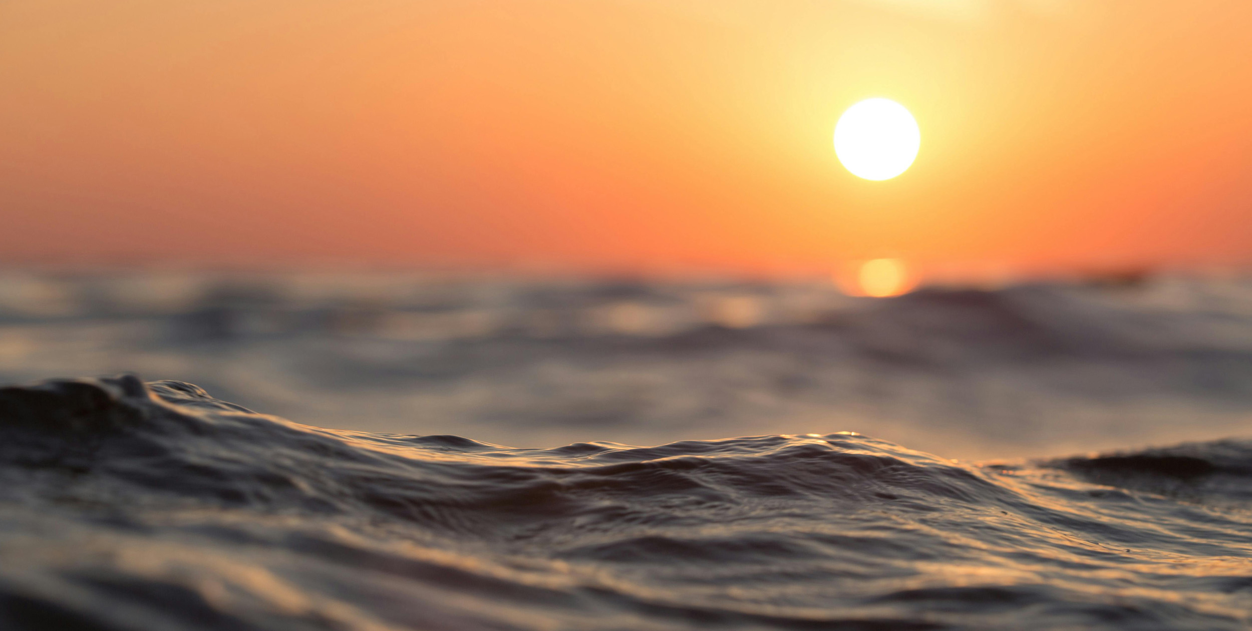 Lugn havsyta i den gyllene timmen. I förgrunden syns små vågor och i bakgrunden en himmel som badar i det varma ljuset från morgonsolen som nyss tagit sig ovanför horisonten.