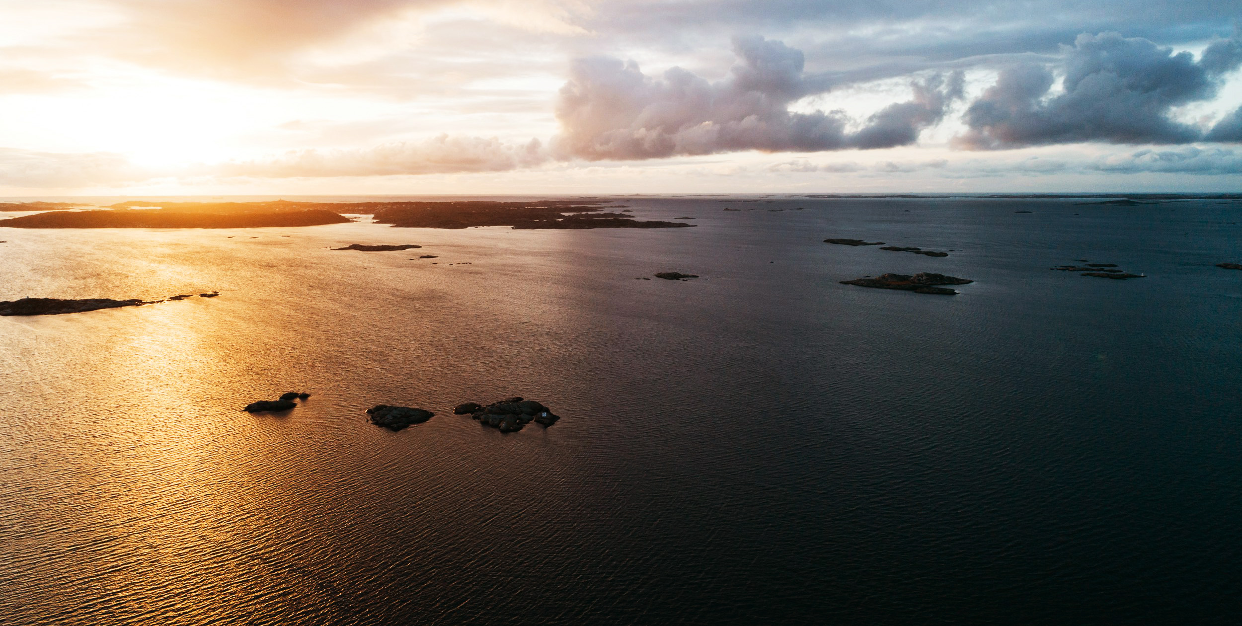 Drönarbild över skärgårdsmiljö. Solen har nyligen gått upp och börjar sprida sitt varma ljus över panoramavyn som visar vatten, kobbar, skär och öar.