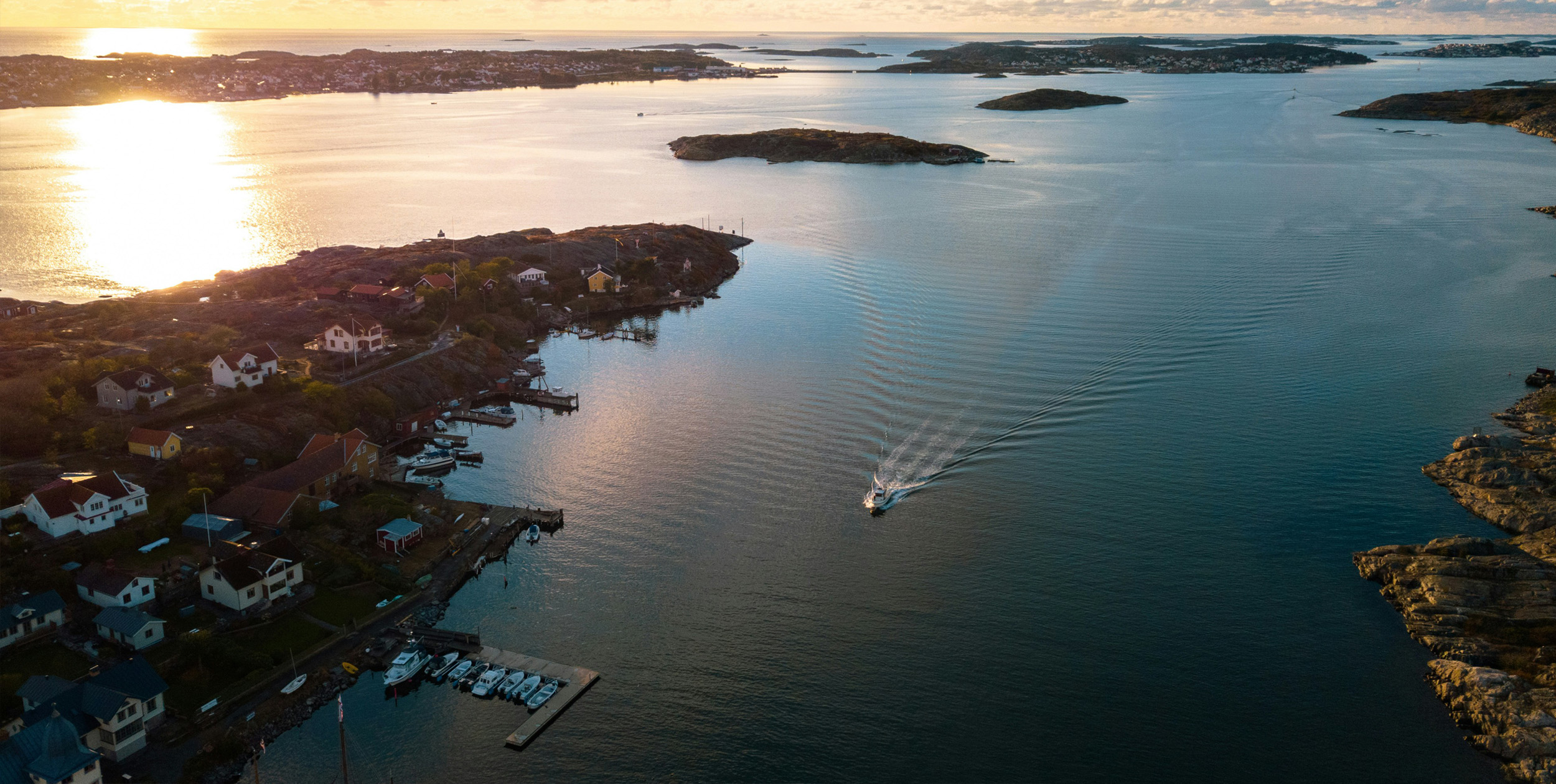 Vy ovanifrån över skärgårdsmiljö i kvällsljus. En ensam båt åker på vattnet. Några hus och båtbryggor med förtöjda båtar syns.