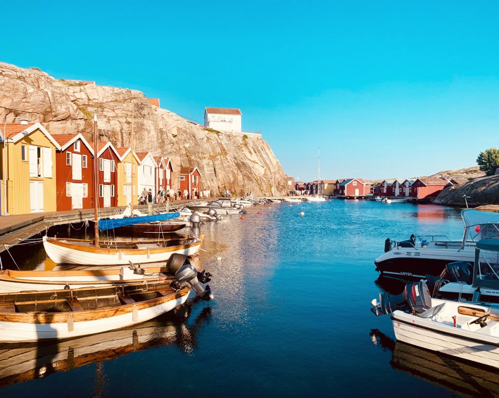 En liten gästhamn där ett antal olika båtar med utombordsmotorer ligger förtöjda. Varmt tidigt kvällsljus mot fiskebodar vid bryggan. Lugnt vatten och klar himmel.