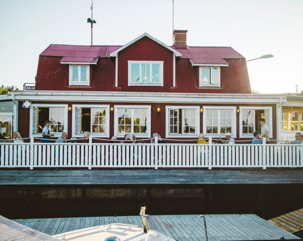Bild på en skärgårdskrog en sommarkväll. I förgrunden skymtar fören på en motorbåt vid bryggan. I bakgrunden syns krogen, ett rött hus med vita knutar och uteservering.